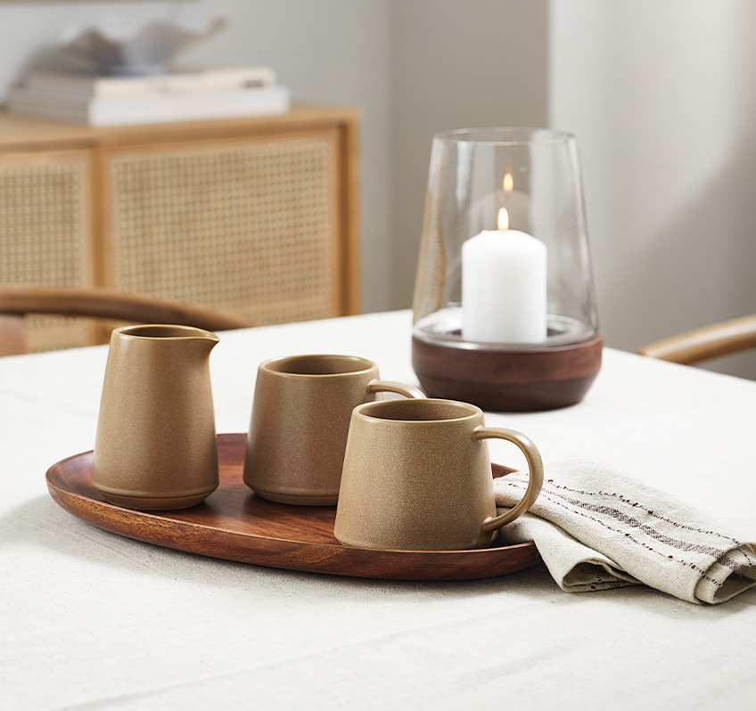 Керамични чаша и каничка за мляко в кафяво на кухненска маса с платнена салфетка и покривка.