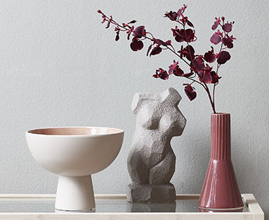 Женска скулптура, купа и розова ваза с изкуствени растения.