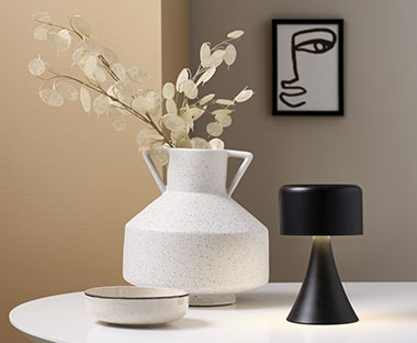 Бяла керамична ваза с изкуствени растения, купа, чврна настолна лампа и рамка за снимка.
