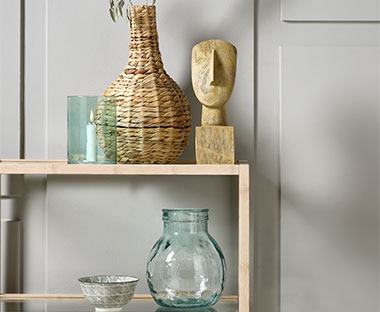 Прозрачна ваза от рециклирано стъкло, плетена ваза, купа, свещник и скулптура върху етажерка.