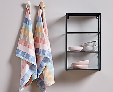 Цветни кухненски кърпи от 100% памук, окачени на стената до миниатюрен рафт с порцеланови купички.