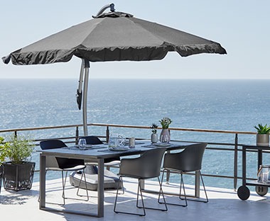 Слънчев балкон с изглед към морето, комплект градински мебели и висящ чадър за слънце.
