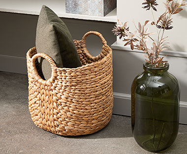 Плетена кошница от воден хиацинт с декоративна възглавница в нея и стъклена ваза.