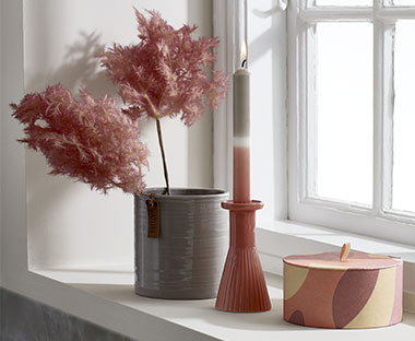 Розова картонена кутия за съхраниение, розов свещник със свещ и ваза с изкуствено растение.