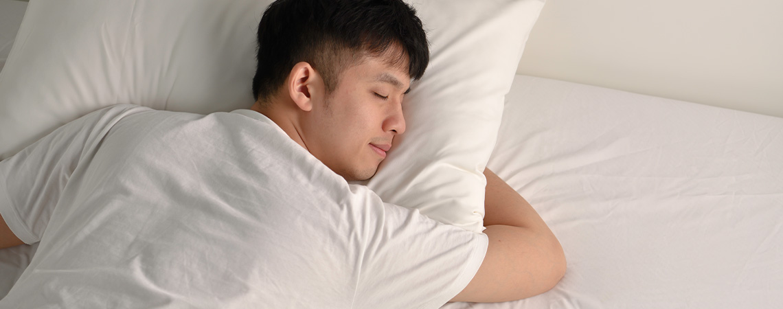 Мъж спи спокойно в легло с бяла възглавница и бял матрак.