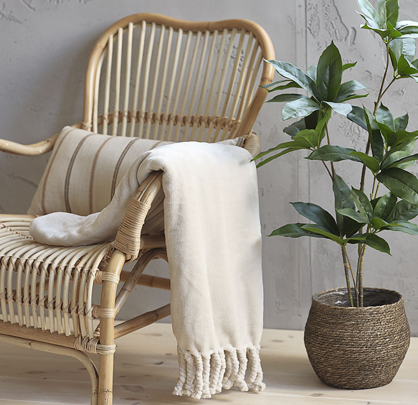 Бежово одеяло върху ратанов стол за отдих и изкуствено растение в саксия.