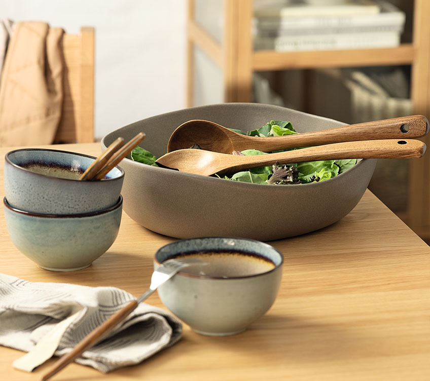 Сива керамична купа за салата с комплект прибори за салата от масивна дървесина върху кухненска маса.